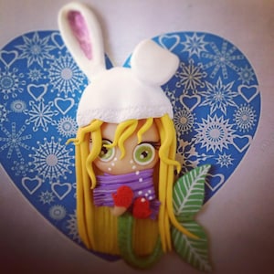 Peut inclure: Figurine fantaisiste d'une fille aux longs cheveux jaunes, portant un chapeau de lapin blanc, une écharpe violette et des mitaines rouges. La figurine a de grands yeux verts et des taches de rousseur. Elle est posée sur un fond bleu en forme de cœur avec des motifs de flocons de neige et de cœurs blancs.