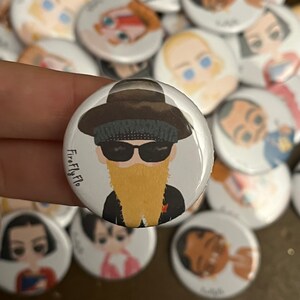 Op de afbeelding: Een witte knop met een cartoonillustratie van een persoon die een zwarte hoed, een zonnebril en een lange gele baard draagt. De tekst "FireFly Flo" staat op de knop gedrukt.