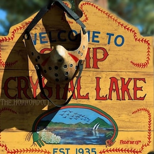 Puede incluir: Un letrero de madera con letras rojas y blancas que dice "Bienvenido al Campamento Crystal Lake Est. 1935". El letrero tiene un borde de puntadas de b&eacute;isbol rojas y blancas. Una m&aacute;scara de hockey blanca con detalles rojos cuelga del letrero.