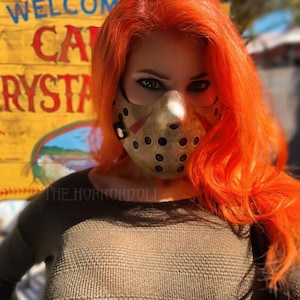 Puede incluir: Una mujer con el pelo naranja brillante lleva una m&aacute;scara de pel&iacute;cula de terror marr&oacute;n y negra. La m&aacute;scara tiene un agujero para la boca y la nariz y est&aacute; cubierta de peque&ntilde;os agujeros. La mujer lleva un su&eacute;ter verde y est&aacute; de pie frente a un cartel amarillo que dice "Bienvenido Camp Crystal".