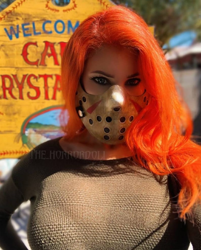 Puede incluir: Una mujer con el pelo naranja brillante lleva una m&aacute;scara de pel&iacute;cula de terror marr&oacute;n y negra. La m&aacute;scara tiene un agujero para la boca y la nariz y est&aacute; cubierta de peque&ntilde;os agujeros. La mujer lleva un su&eacute;ter verde y est&aacute; de pie frente a un cartel amarillo que dice "Bienvenido Camp Crystal".