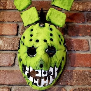 May include: A lime green mask with black eye holes and black dots. The mask has a wide, toothy grin with white teeth. Four green, fluffy rectangles are attached to the top with wire. A black spike is on top.