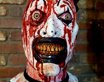 May include: A grotesque clown mask with a white face, red blood-like splatters, and a black top hat. The mask features exaggerated features, including a wide, menacing grin with yellow teeth and black lipstick. The clown's ruffled collar is also stained with red.