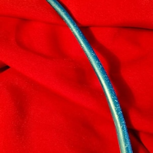 Puede incluir: Una varita azul curvada con motas blancas, sobre un fondo rojo vibrante. La varita tiene un acabado liso y brillante y una punta ligeramente puntiaguda. Parece estar hecha de un material sólido.
