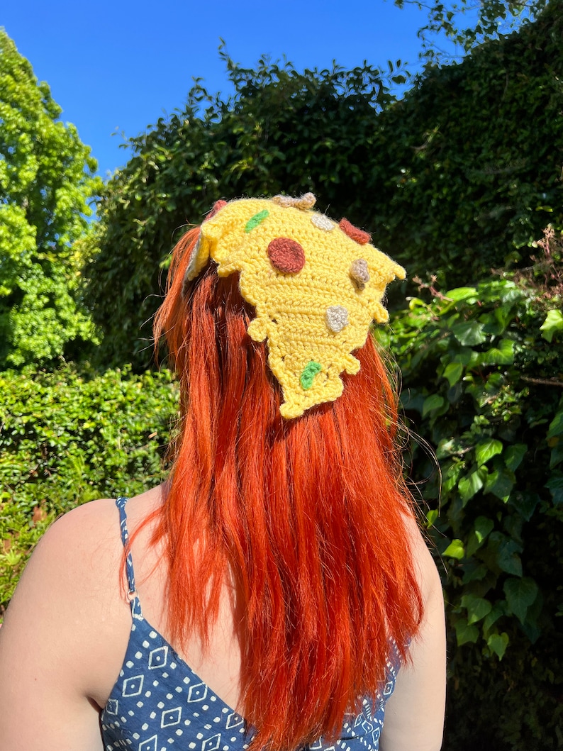 Puede incluir: Una persona con el pelo largo y rojo lleva una diadema de pizza hecha a crochet. La diadema es amarilla con detalles verdes, marrones y blancos.