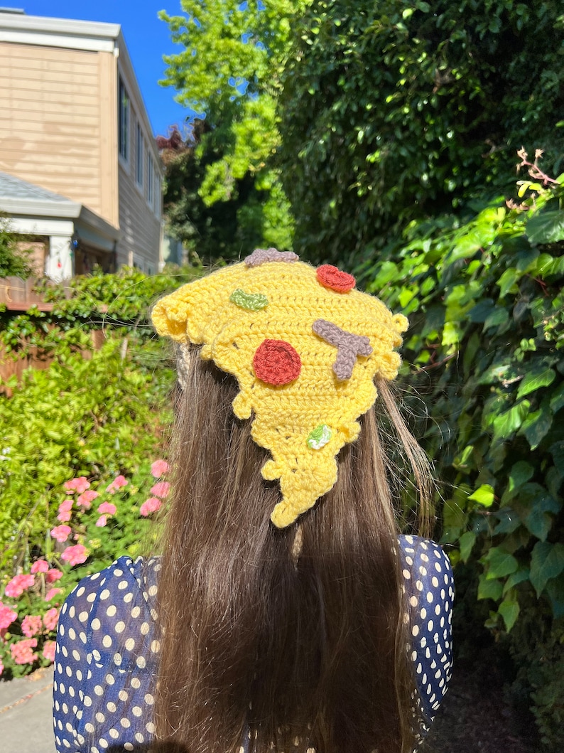 Puede incluir: Un gorro de pizza de crochet amarillo con ingredientes rojos, verdes y marrones. El gorro lo lleva una persona con el pelo largo y casta&ntilde;o y una camisa de lunares azules y blancos.