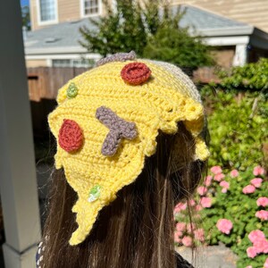 Puede incluir: Un gorro de pizza de crochet amarillo con ingredientes rojos, verdes y marrones. El gorro lo lleva una persona con el pelo largo y casta&ntilde;o.