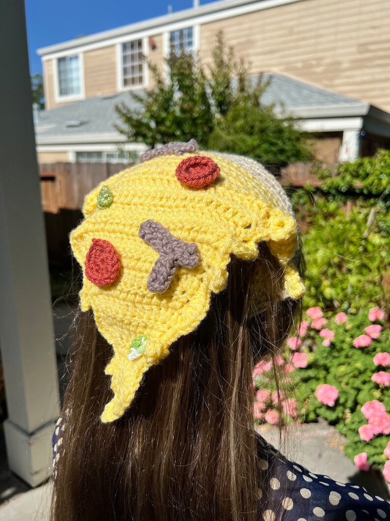 Puede incluir: Un gorro de pizza de crochet amarillo con ingredientes rojos, verdes y marrones. El gorro lo lleva una persona con el pelo largo y casta&ntilde;o.