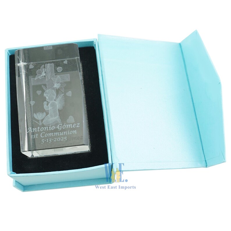 May include: A clear glass cube with an engraved image of an angel with wings, a cross, and butterflies. The cube is in a light blue box with the text "Antonio Gomez 1st Communion 5-13-2025" engraved on the front.