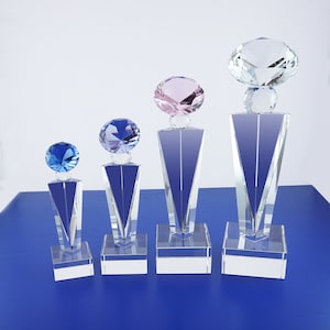 May include: Four crystal award trophies with different colored diamond tops. The trophies are arranged in order of size, with the largest trophy on the right and the smallest on the left. The diamond tops are clear, pink, blue, and light blue.