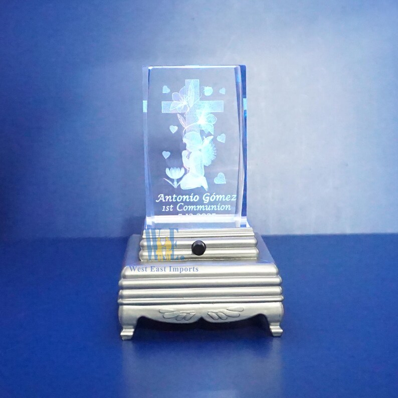 May include: A clear glass cube with an engraved image of an angel holding a cross, surrounded by hearts. The cube is on a silver base with the text "Antonio Gomez 1st Communion" and "West East Imports".