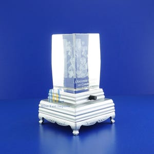 May include: A clear glass cube with an engraved image of a cross and angels. The cube is on a silver base with a music box mechanism. The base has the text "West-East Imports" and "J. Antonio 1st Communion 5-12-2013".