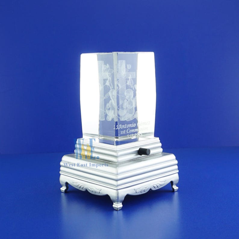 May include: A clear glass cube with an engraved image of a cross and angels. The cube is on a silver base with a music box mechanism. The base has the text "West-East Imports" and "J. Antonio 1st Communion 5-12-2013".