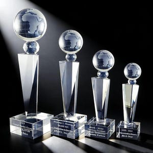 May include: Four clear crystal trophies of different sizes, each with a globe design on top of a faceted pedestal. The bases have engraved text with names, dates, and titles. The trophies are set against a dark background.