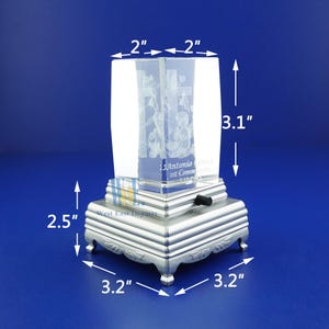 May include: A clear glass cube with a 3D engraved image of a cross and flowers. The cube is mounted on a silver metal base with a wind-up music box mechanism. The base measures 3.2 inches by 3.2 inches and is 2.5 inches tall. The cube is 2 inches by 2 inches and 3.1 inches tall.