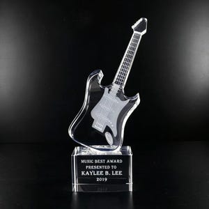 May include: A clear acrylic guitar-shaped award on a rectangular base. The guitar has etched details and is mounted on a clear base with the text "MUSIC BEST AWARD PRESENTED TO KAYLEE B. LEE 2019".