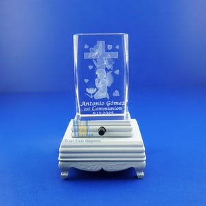 May include: A clear crystal cube with an engraved image of an angel kneeling before a cross with flowers. The cube is on a white wooden base with the text "Antonio Gomez 1st Communion 5-13-2025" and "West East Imports".