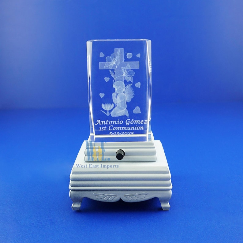May include: A clear crystal cube with an engraved image of an angel kneeling before a cross with flowers. The cube is on a white wooden base with the text "Antonio Gomez 1st Communion 5-13-2025" and "West East Imports".