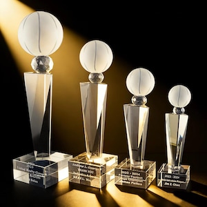 Puede incluir: Cuatro trofeos de cristal transparente de diferentes tamaños. Cada trofeo presenta una esfera esmerilada en la parte superior, una base de cristal facetado y una base rectangular con texto grabado. El texto incluye nombres, fechas e información de la empresa.