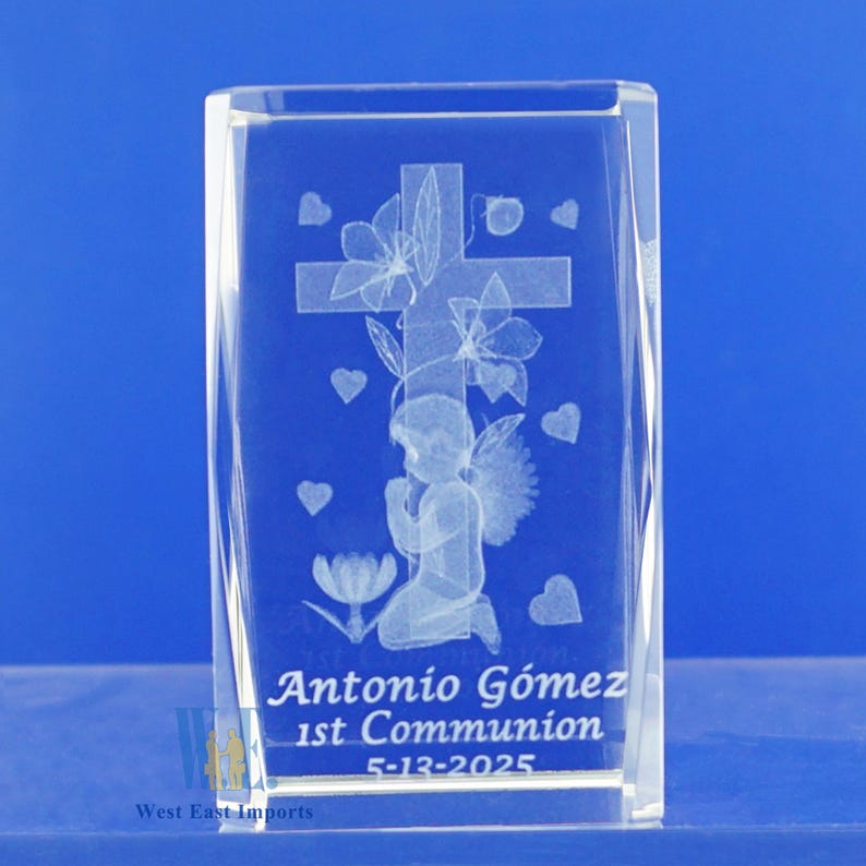 May include: A clear glass cube with an engraved image of a praying angel with wings, a cross, flowers, and hearts. The text "Antonio G&oacute;mez 1st Communion 5-13-2025" is engraved at the bottom.