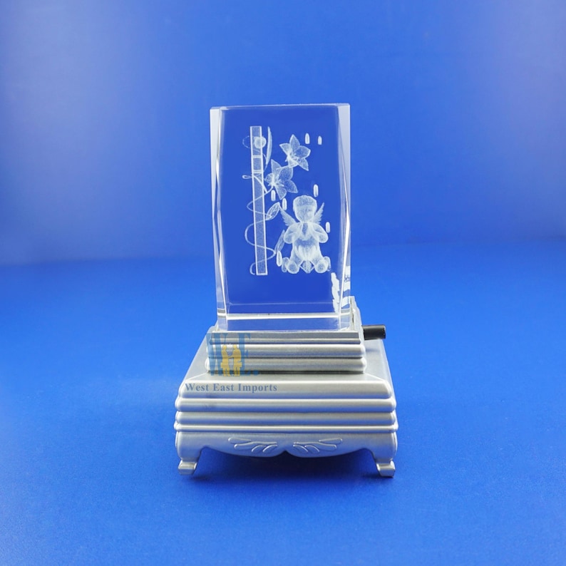May include: A clear glass cube with a 3D etched design of an angel with wings and flowers. The cube is on a silver base with the words "West East Imports" printed on it.