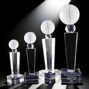 May include: Four clear crystal trophies of varying heights, each featuring a frosted glass golf ball topper on a faceted crystal sphere, supported by a tapered rectangular column and a square base. The tallest trophy base is engraved with text.