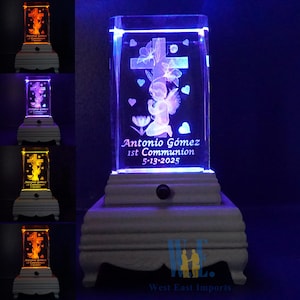 Puede incluir: Una base de madera blanca con un cubo de cristal transparente en la parte superior. El cubo tiene una imagen grabada en 3D con láser de un ángel arrodillado en oración con una cruz detrás. La base tiene un interruptor de luz en el lateral y las palabras "Antonio Gómez 1ª Comunión 5-13-2025" están grabadas en el cubo de cristal.