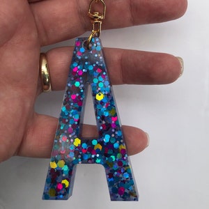 May include: A blue resin letter A keychain with iridescent glitter. The letter is attached to a gold metal key ring.