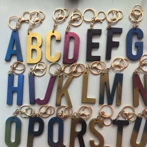 May include: A set of 26 alphabet letter keychains. Each letter is made of wood and has a different color gradient. The keychains are attached to gold-colored metal rings.