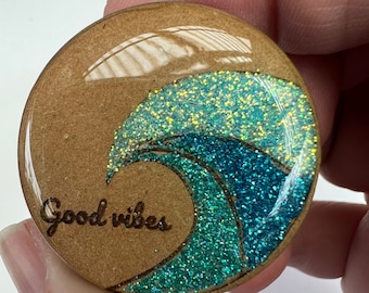May include: A round wooden badge holder with a blue and green glitter wave design and the text "Good vibes" engraved on the front.