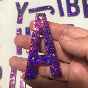 May include: A glittery purple and pink letter A with a hole at the top. The letter is made of resin and is covered in iridescent glitter.