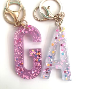 May include: Two gold keychains with a pink and white letter charm. The letters are "G" and "A" and are filled with colorful glitter.