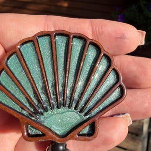 May include: A seashell-shaped badge reel featuring a dark brown wooden frame and a shimmering, glittery mint green resin inlay. The design includes distinct vertical ridges radiating from the base of the shell, attached to a black plastic clip.