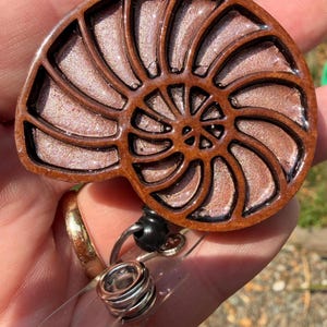 May include: A badge reel featuring a nautilus shell design with a dark brown frame and a shimmering, metallic rose gold interior. The circular shell is attached to a retractable black cord mechanism with a clear plastic strap and metal snap.