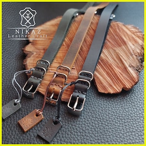 Pu&ograve; includere: Tre cinturini per orologi in pelle grigia, marrone e nera, ciascuno con una fibbia argentata e un piccolo cartellino. I cinturini sono esposti su una superficie di legno, mettendo in risalto la maestria artigianale e i materiali.