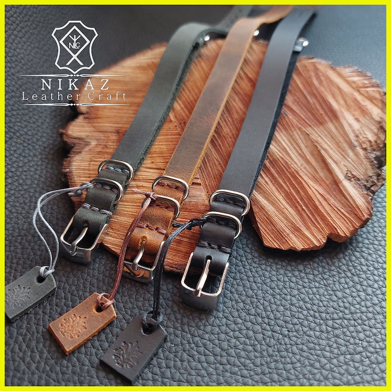 Pu&ograve; includere: Tre cinturini per orologi in pelle grigia, marrone e nera, ciascuno con una fibbia argentata e un piccolo cartellino. I cinturini sono esposti su una superficie di legno, mettendo in risalto la maestria artigianale e i materiali.