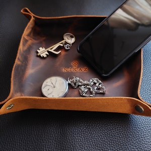 May include: A brown leather tray with a key, a pocket watch, a silver chain, and a smartphone. The tray has the word "NIKAZ" stamped on it. The tray is rectangular with rounded corners.