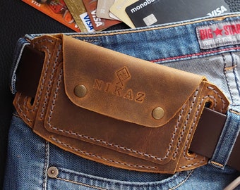 May include: A brown leather belt bag with the word "NIKAZ" embossed on the front. The bag is attached to a brown belt and is resting on blue jeans. Several credit cards are visible in the background.