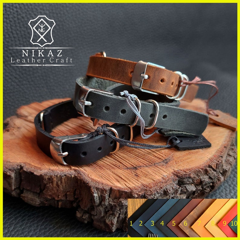Pu&ograve; includere: Tre braccialetti in pelle marrone, nera e grigia, ciascuno con una fibbia argentata. I braccialetti sono esposti su una superficie di legno. Il logo del marchio "NIKAZ Leather Craft" &egrave; visibile in alto a sinistra.