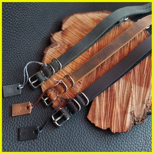 Pu&ograve; includere: Tre cinturini per orologi in pelle nera, marrone e grigia, ciascuno con una fibbia argentata e un piccolo cartellino. I cinturini sono esposti su una superficie di legno con uno sfondo in pelle scura.