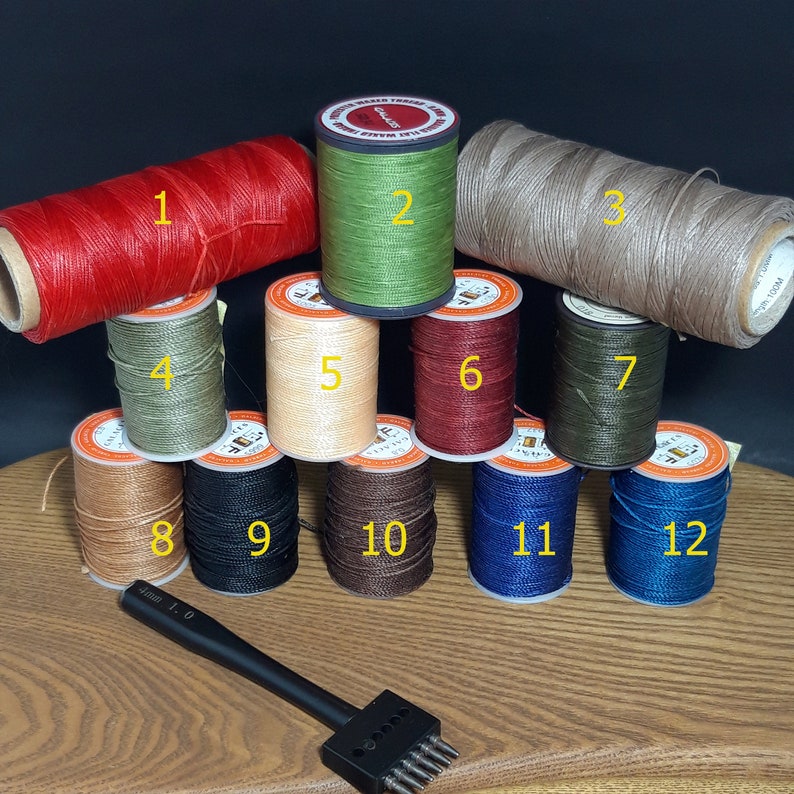Pu&ograve; includere: Una collezione di rocchetti di filo cerato in vari colori, tra cui rosso, verde, beige e blu. &Egrave; presente anche un punzone per lavori in pelle nero. Le rocchetti sono disposti su una superficie di legno.