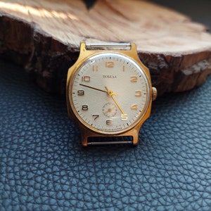 May include: A vintage gold-coloured wristwatch with a cream face. The watch has black numerals, gold hands, and the word "ПОБЕДА" in black. It is set on a black leather surface.