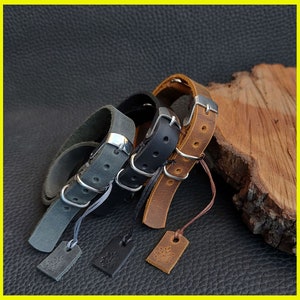 Pu&ograve; includere: Braccialetti in pelle grigia, nera e marrone con fibbie argentate ed etichette attaccate. I braccialetti sono esposti su una superficie scura, mettendo in risalto la loro consistenza e il loro design. Ogni braccialetto ha un colore diverso e un'etichetta unica.