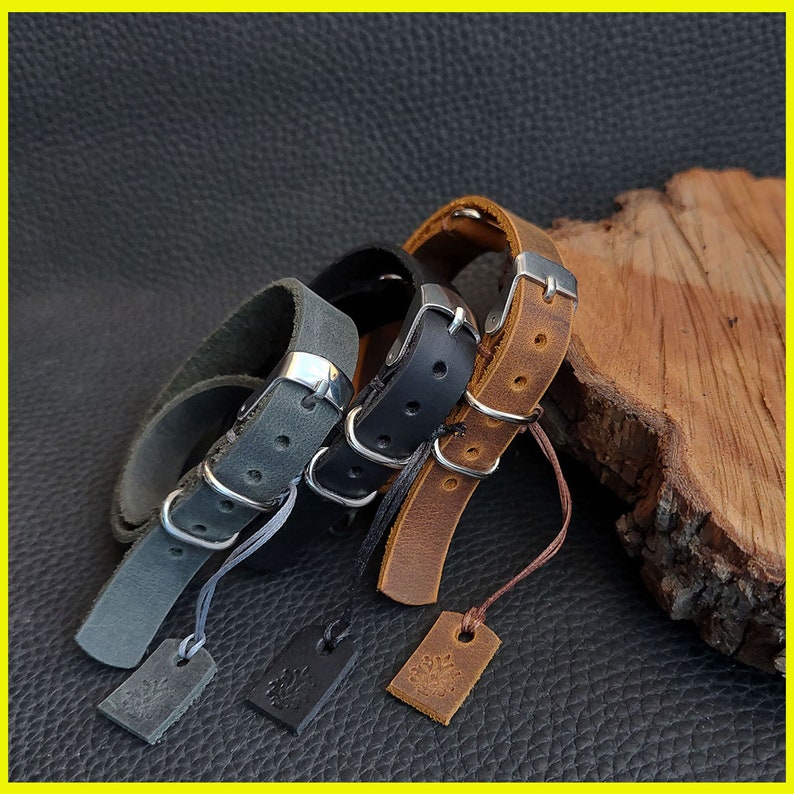 Pu&ograve; includere: Braccialetti in pelle grigia, nera e marrone con fibbie argentate ed etichette attaccate. I braccialetti sono esposti su una superficie scura, mettendo in risalto la loro consistenza e il loro design. Ogni braccialetto ha un colore diverso e un'etichetta unica.