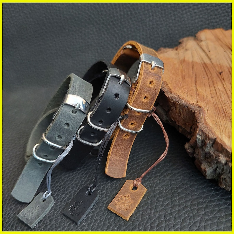 Pu&ograve; includere: Tre braccialetti in pelle grigia, nera e marrone, ciascuno con una fibbia argentata e un piccolo tag. I braccialetti sono esposti su una superficie scura, mostrando la loro consistenza e il design.