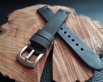 May include: Black leather watch straps with a silver-colored buckle. The straps are displayed on a wooden surface. The watch straps have multiple holes for adjustment.