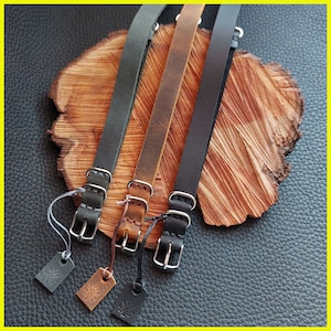 Pu&ograve; includere: Tre cinturini per orologi in pelle nera, marrone e verde scuro, ciascuno con una fibbia argentata e una piccola etichetta. I cinturini sono esposti su una superficie di legno con uno sfondo in pelle strutturata.
