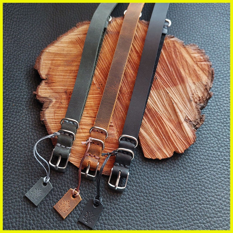 Pu&ograve; includere: Tre cinturini per orologi in pelle nera, marrone e verde scuro, ciascuno con una fibbia argentata e una piccola etichetta. I cinturini sono esposti su una superficie di legno con uno sfondo in pelle strutturata.