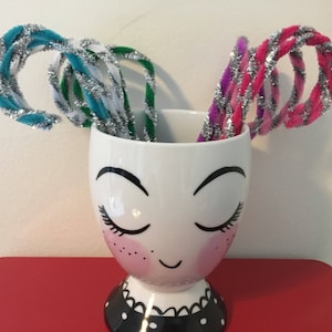 May include: A white ceramic mug with a painted face and eyelashes, decorated with a black and white polka dot pattern around the base. The mug is filled with colorful pipe cleaners.