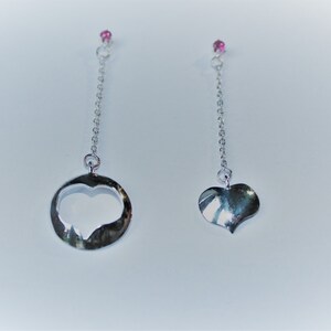 Könnte beinhalten: Ein Paar silberne Ohrringe. Ein Ohrring hat einen herzförmigen Anhänger in einem Kreis, der andere ein massives Herz. Beide hängen an einer Kette und sind mit einem kleinen rosa Edelstein versehen.